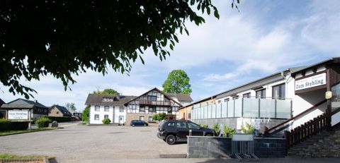 Ein ruhiger Parkplatz vor einem Hotel mit mehreren Gebäuden. Der Himmel ist klar und sonnig, umgeben von Bäumen.