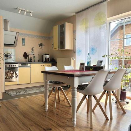 Een lichte keuken met een eettafel en stoelen. De grote ramen laten veel daglicht binnen en er zijn enkele planten in de kamer.