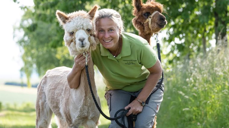 A person stands in nature and smiles while holding two alpacas on leashes. In the background, trees and suspicious grass are visible.