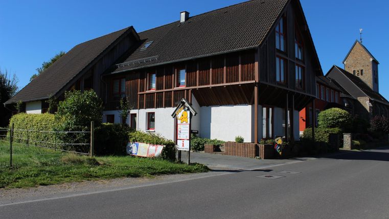 Ein modernes Haus mit Holzverkleidung, umgeben von grünem Rasen und Bäumen. Im Hintergrund ist ein Kirchturm sichtbar und der Himmel ist klar und blau.