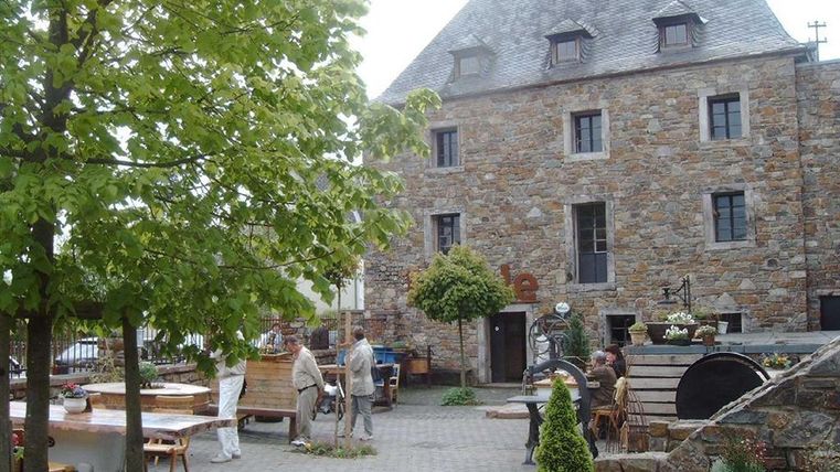 Ein historisches Steinhaus mit mehreren Fenstern und einem charakteristischen Dach. Umgeben von einem gemütlichen Innenhof mit Bäumen und Tischen.