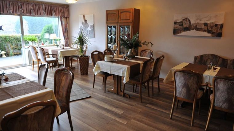 A cozy restaurant with wooden furniture and beautifully set tables. Large windows offer a view of the outdoor area.