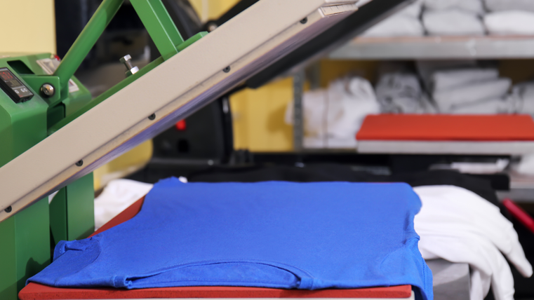 Ein blaues T-Shirt liegt in einer Stoffpresse. Im Hintergrund sieht man ein Reagl mit gefalteten weißen T-Shirts.