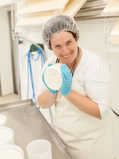 Eine lächelnde Frau trägt Handschuhe und hält einen Behälter in der Hand. Im Hintergrund sind weitere Behälter zu sehen, die auf einer Arbeitsfläche stehen.
