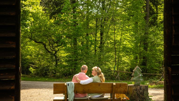 Ein Paar sitzt auf einer Bank im Wald und genießt die Natur. Um sie herum sind grüne Bäume und ein ruhiger Weg.