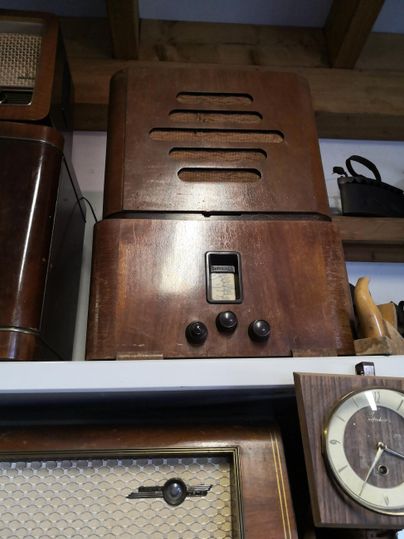 Ein vintage Holzradio mit drei Knöpfen und einem Lüftungsgitter. Es steht auf einem Regal und ist Teil einer Sammlung alter Geräte.