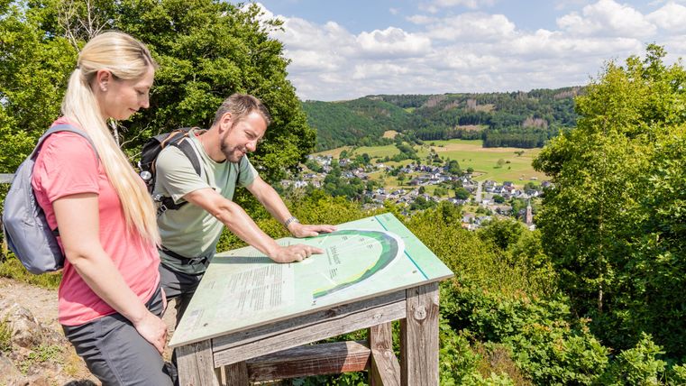 Two hikers are looking at a map at a viewpoint. In the background, a beautiful landscape with hills and a small village can be seen.