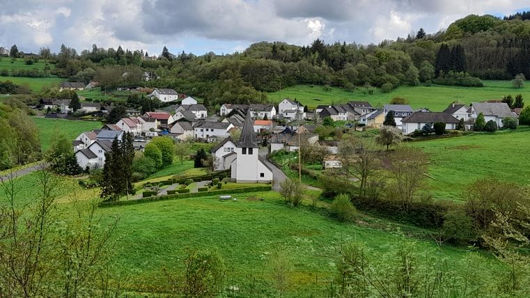 A picturesque village in a green landscape. The houses are surrounded by meadows and trees, with a small church in the foreground.