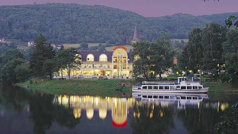 Ein malerischer Blick auf ein Gebäude am Fluss mit sanften Hügeln im Hintergrund. Auf dem Wasser spiegelt sich das Licht des Gebäudes.
