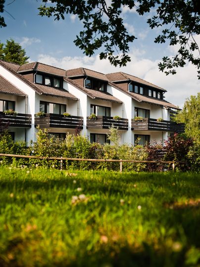 Een modern gebouw met balkons, omringd door groen gras en bomen. De lucht is gedeeltelijk bewolkt, wat een uitnodigende sfeer creëert.