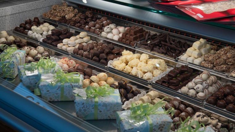 Eine ansprechende Auswahl an Pralinen und Schokoladen in einem Vitrinenauslage. Klein verpackte Geschenke mit bunten Schleifen sind ebenfalls sichtbar.