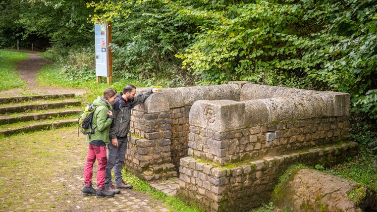 Zwei Personen betrachten einen historischen Brunnen in einer Waldumgebung.