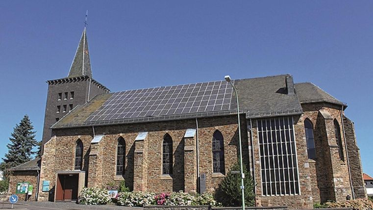 A church with a steep spire and solar panels on the roof. The facade is made of stone and the windows are designed in Gothic style.