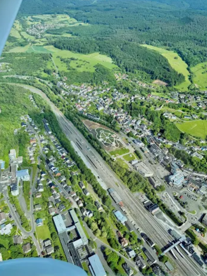 Luchtfoto van een plek met een station, woonwijken en veel groen. Een ronde bloemenperkje is zichtbaar naast de sporen.
