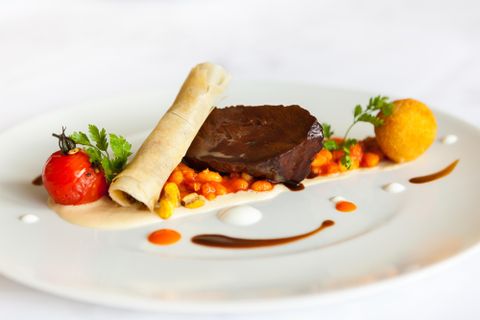 Ein kunstvoll angerichteter Teller mit saftigem Fleisch, serviert mit einer Beilage aus buntem Gemüse und einer Sauce. Kleine Tomaten und Kräuter ergänzen die Präsentation.