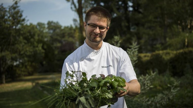 Een kok staat buiten en houdt verse kruiden in zijn hand. Op de achtergrond zijn bomen en een groene landschap te zien.