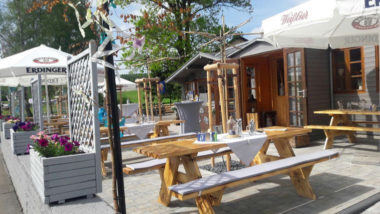 Eine einladende Außenterrasse mit Holztischen und bunten Blumen. Über den Tischen hängen Sonnenschirme und es gibt eine rustikale Holzhütte im Hintergrund.