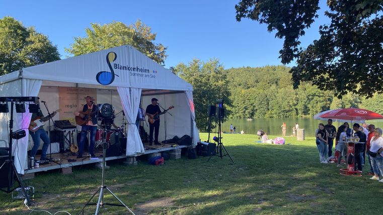 A band is playing live under a tent by the shore of a lake. In the background, there are people relaxing and enjoying nature.