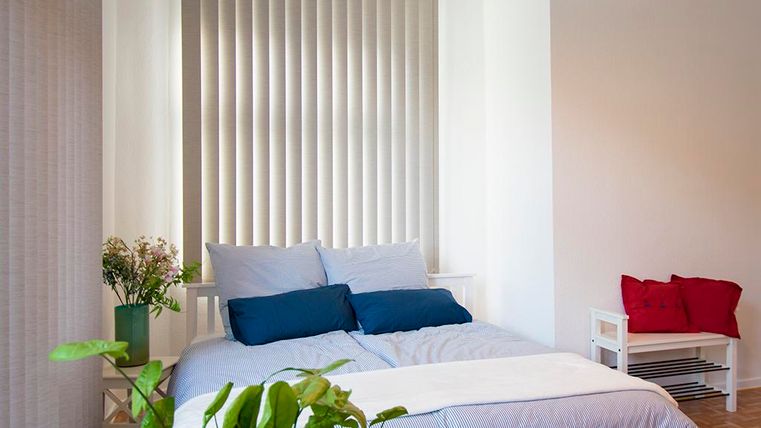 Ein helles Schlafzimmer mit einem Bett, das blaue und weiße Kissen hat. Pflanzen und ein kleiner Tisch ergänzen die gemütliche Atmosphäre.