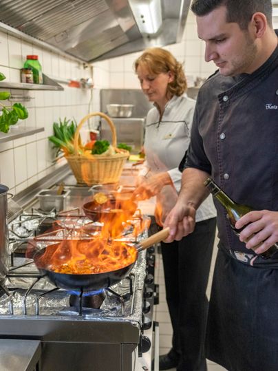 In a modern kitchen, a chef is cooking with a flaming pan's contents. A woman is smiling at him while fresh vegetables are visible in the background.