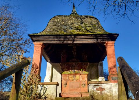 Eine kleine Kapelle mit einer markanten Dachform steht auf einem Hügel. Die Umgebung ist von Bäumen umgeben und der Himmel ist blau.