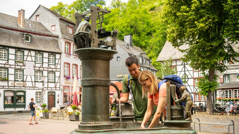 Zwei Personen erfrischen sich am Tuchmacherbrunnen in Monschau, umgeben von Fachwerkhäusern und Bäumen.