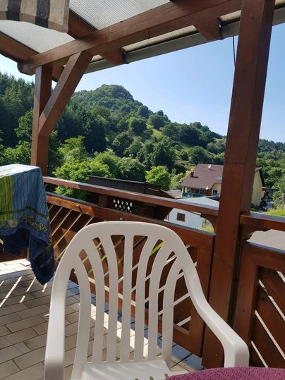 Een balkon met een witte stoel en uitzicht op groene heuvels. De lucht is helder en zonnig, wat een ontspannen sfeer creëert.