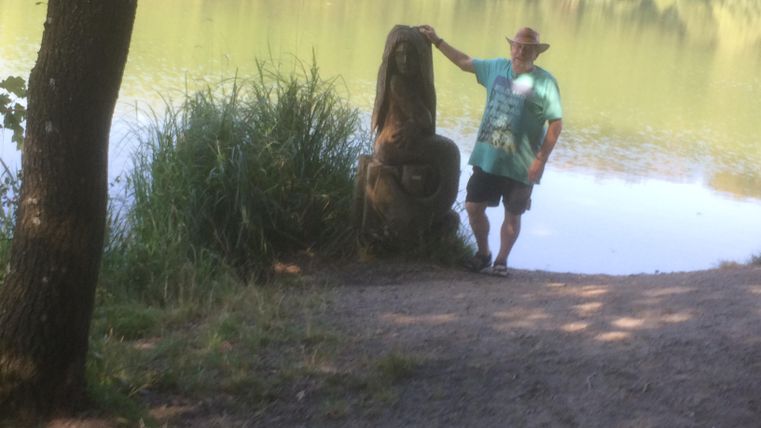 Ein Mann steht neben einer Statue am Ufer eines ruhigen Sees. Die Umgebung ist grün und bewaldet, mit einer klaren Wasseroberfläche.