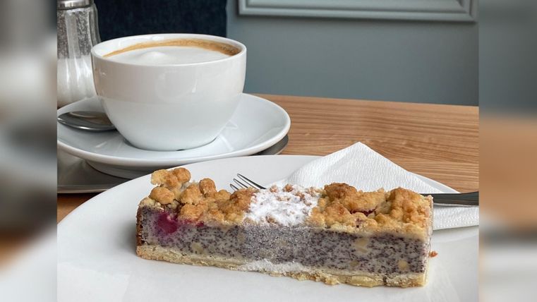 Een stuk taart met een kruimelige bovenkant ligt op een wit bord. Naast het bord staat een kopje koffie op een schoteltje.