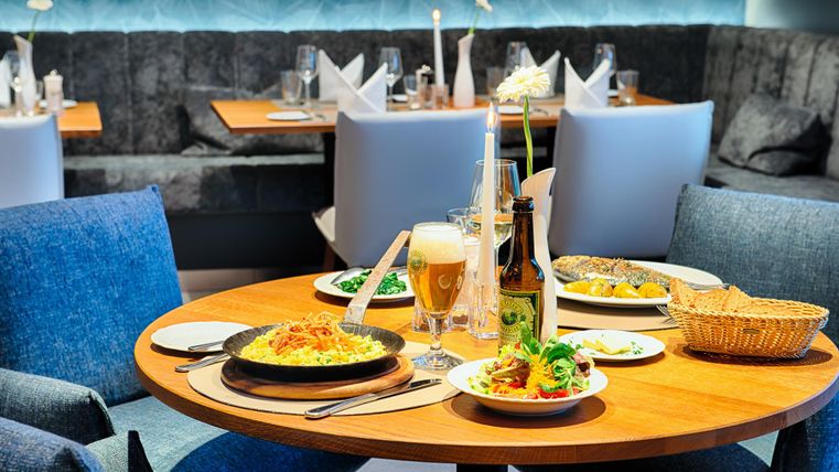 Een tafel in een restaurant met verschillende gerechten, waaronder pasta, salade en vis. Een bierglas en een fles staan ernaast.