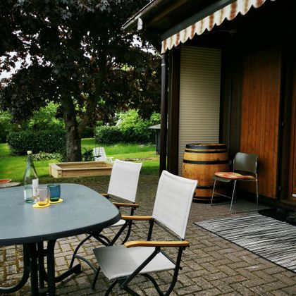 Een gezellige buitenruimte met een tafel en stoelen. Op de achtergrond ziet men een goed onderhouden tuin met groene vegetatie.