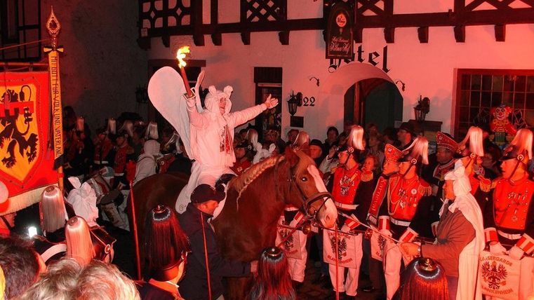 A festive event featuring a costumed rider on a horse. In the background, people in historical uniforms and torches can be seen.