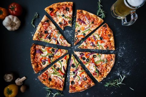 Pizza in eight slices with vegetables and olives, surrounded by tomatoes, garlic, and a beer glass on a black background.