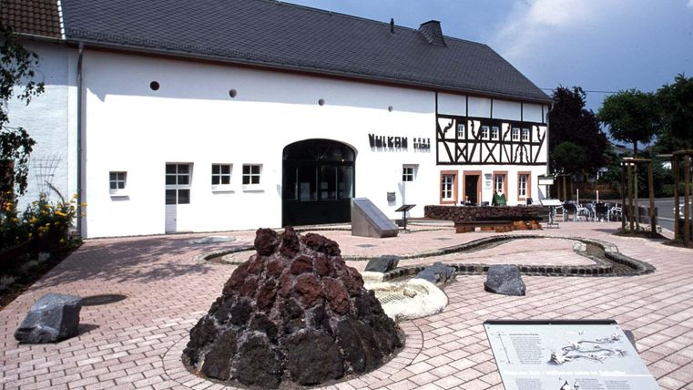 Ein historisches Gebäude mit einem modernen Eingang und einer malerischen Umgebung. Vor dem Gebäude befindet sich eine künstlerische Landschaftsgestaltung mit Steinen und Vulkanformationen.