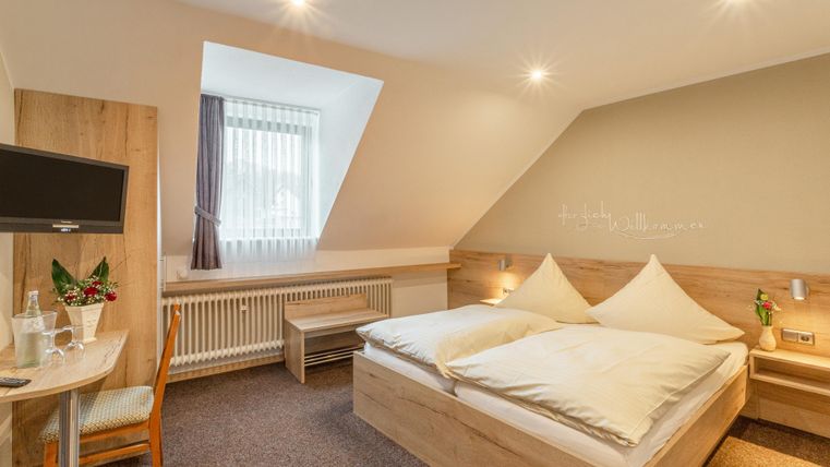 Een gezellige hotelkamer met een tweepersoonsbed en een klein tafeltje. De muren zijn licht en er komt veel daglicht binnen door het raam.