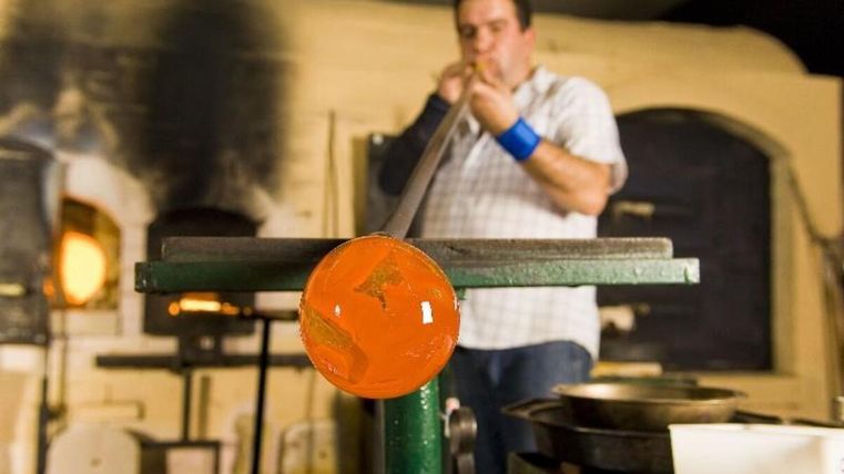 Ein Glasbläser arbeitet an einem Glasstück in einer Werkstatt. Im Hintergrund sind Öfen und Werkzeuge zu sehen.