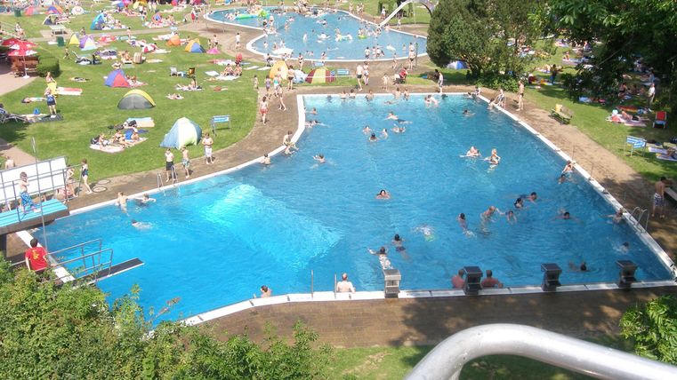 Ein großer Swimmingpool mit vielen Schwimmern und Freizeitaktivitäten. Rundherum sind Liegewiesen und Sonnenschirme aufgestellt.