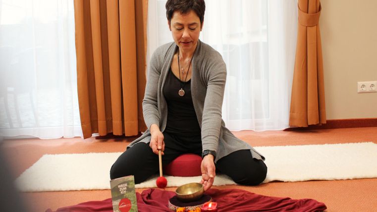 Eine Frau sitzt im Schneidersitz auf dem Boden und spielt mit einer Klangschale. Vor ihr liegt ein rotes Tuch mit verschiedenen Gegenständen.