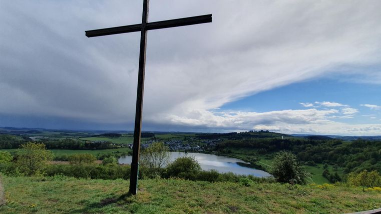 Ein großes Kreuz steht auf einem Hügel mit Blick auf eine ruhige Landschaft. Im Hintergrund sind ein See und ein weitläufiger, bewölkter Himmel zu sehen.