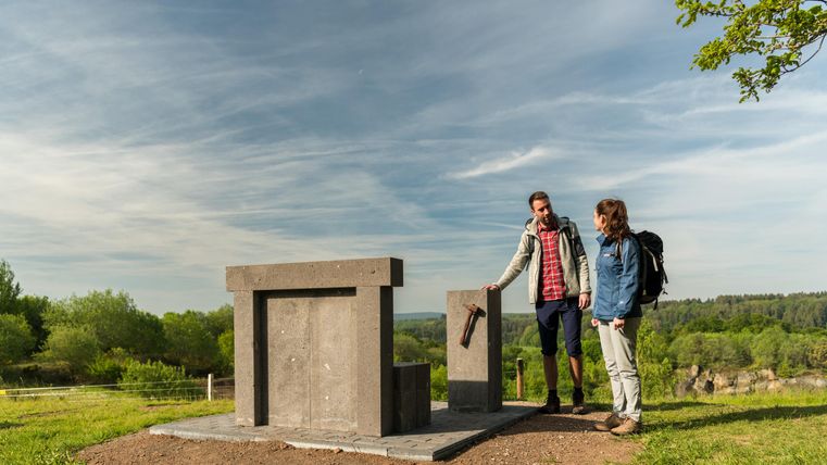 Zwei Personen betrachten eine steinerne Skulptur im Freien. Die Landschaft ist grün und der Himmel leicht bewölkt.