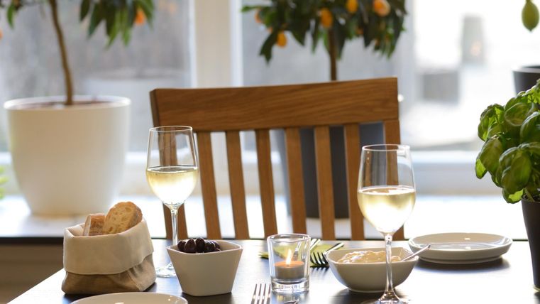 Ein gedeckter Tisch mit zwei Gläsern Weißwein und verschiedenen kleinen Schalen. Im Hintergrund stehen Pflanzen mit orangefarbenen Früchten.