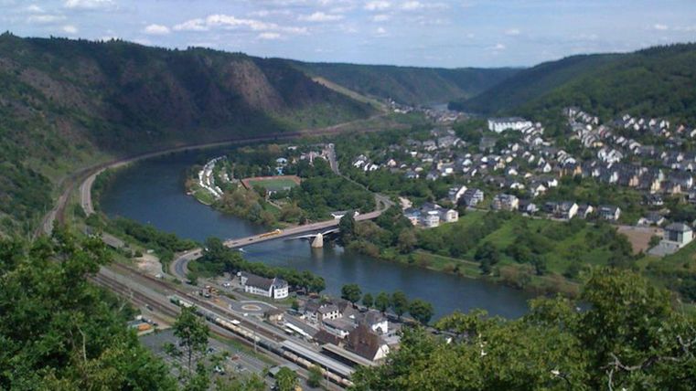 Een schilderachtig landschap met een rivier die door groene heuvels stroomt. Op de achtergrond zijn kleine huizen en een brug te zien.