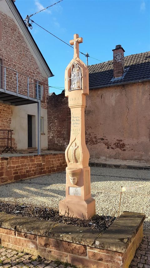 Een steen kruis staat in een kleine nederzetting. De lucht is blauw en de omgeving lijkt rustig en landelijk.