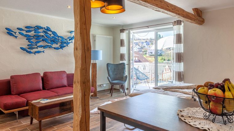 Ein gemütlicher Wohnraum mit rotem Sofa und blauer Wanddekoration. Der Raum hat große Fenster mit Blick auf den Balkon und eine Obstschale auf dem Tisch.