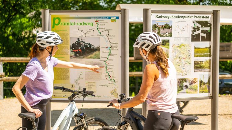 Twee fietsters in helmen kijken naar een kaart van de Prüm-vallei fietsroute. Ze plannen hun route en lijken te discussiëren over het verloop van de weg.