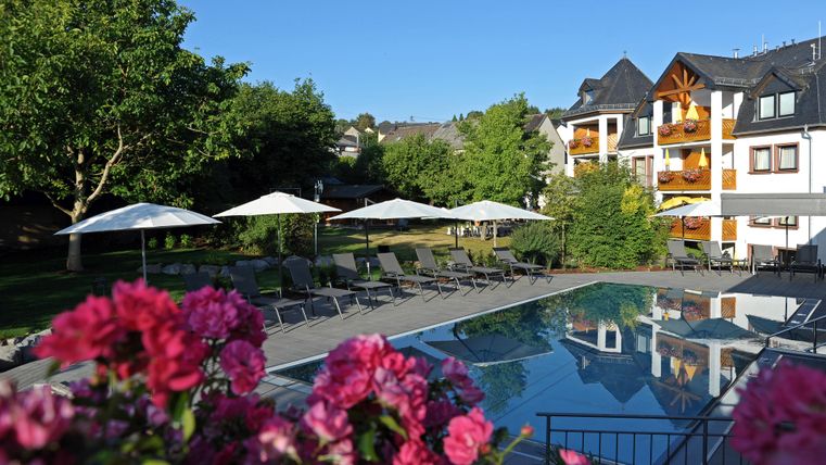 Ein entspannender Poolbereich mit Liegen und Sonnenschirmen. Im Hintergrund ist ein schönes Hotelgebäude von üppigem Grün umgeben.