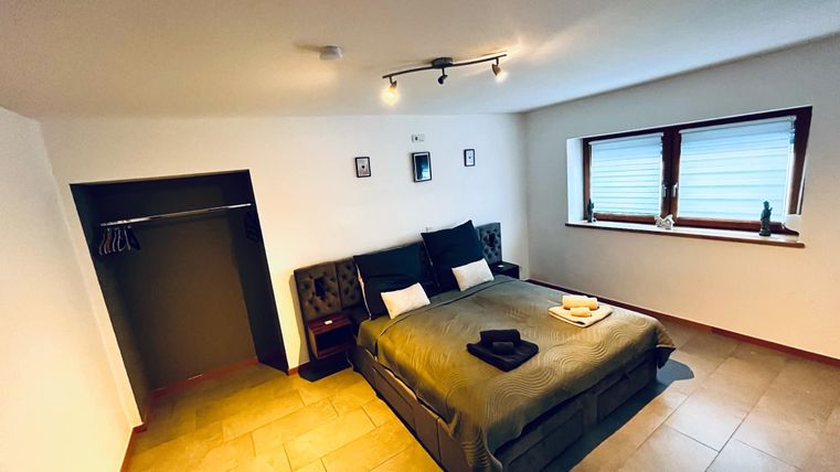 A modernly furnished bedroom with a large double bed and a simple design. There is a window with blinds and a wardrobe.