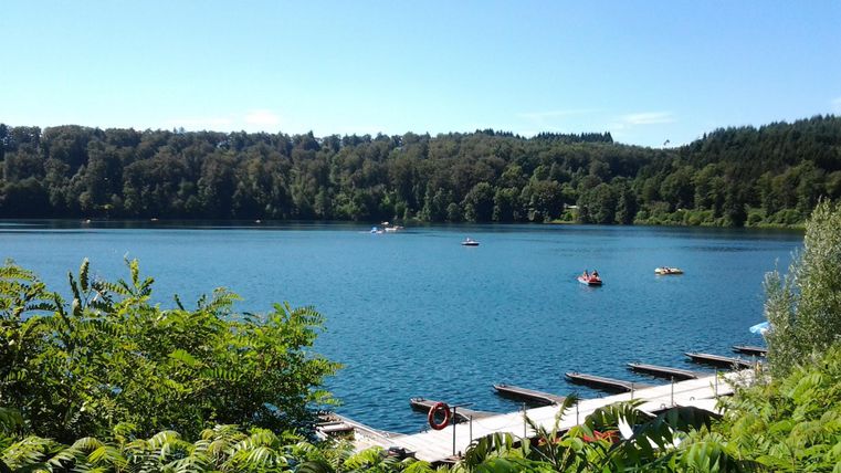 Ein ruhiger See umgeben von Bäumen. Kleine Boote schwimmen auf dem Wasser, und ein Steg führt ins Wasser.