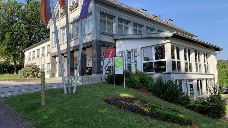 A modern building with many windows and a well-kept garden. In front of the house, there are flags and colorful flower beds.