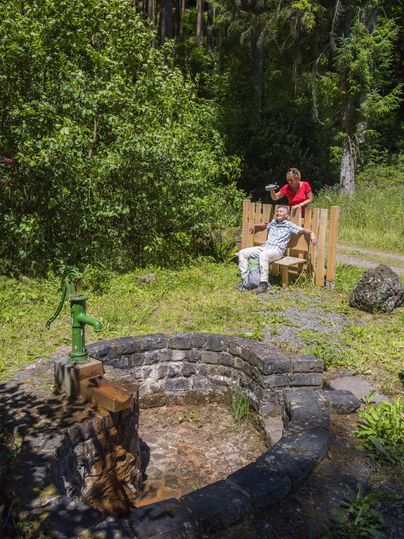 Eine Person sitzt auf einer Bank in der Natur, während eine andere Person am Brunnen steht. Umgeben von Bäumen und grünem Gras vermittelt die Szenerie eine ruhige Atmosphäre.